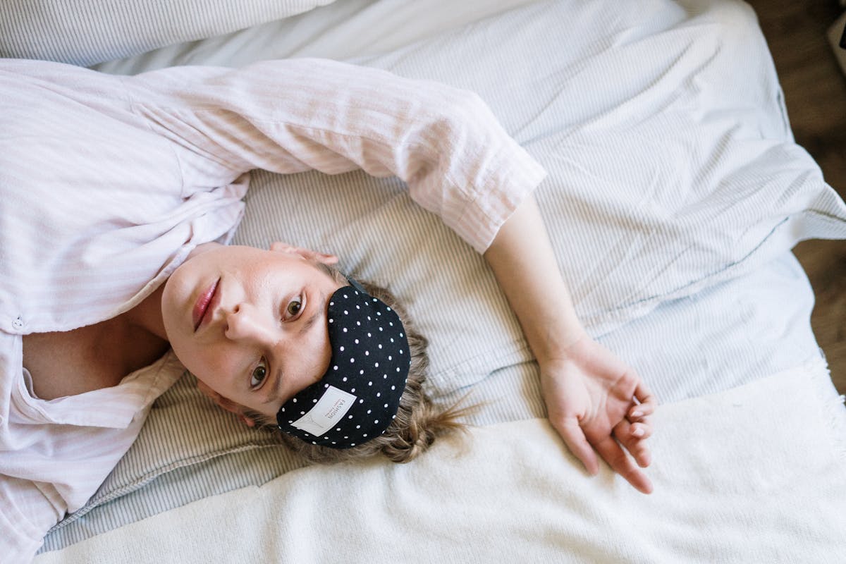 Woman lying on a bed with a sleep mask partially covering her forehead, looking relaxed and resting.